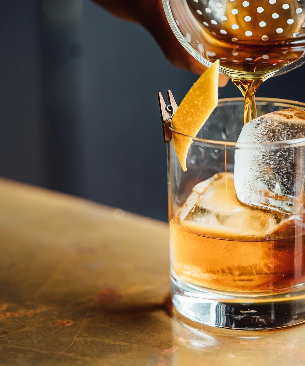 A cocktail being poured over ice on a worn brass bar